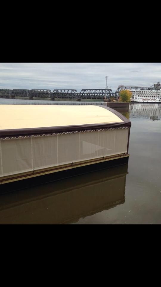 Floating enclosed structure with tan roof, water, distant bridge and buildings under overcast sky.