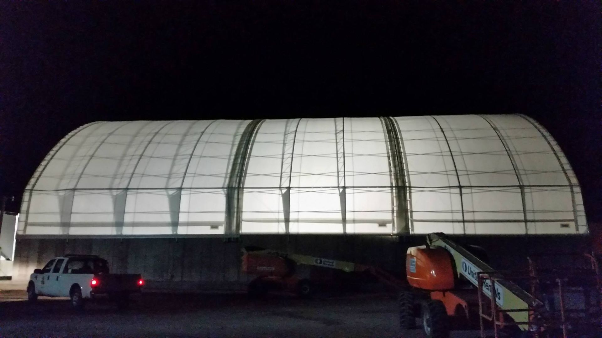Large, illuminated white arched-roof building at night, with a truck and lift in front.