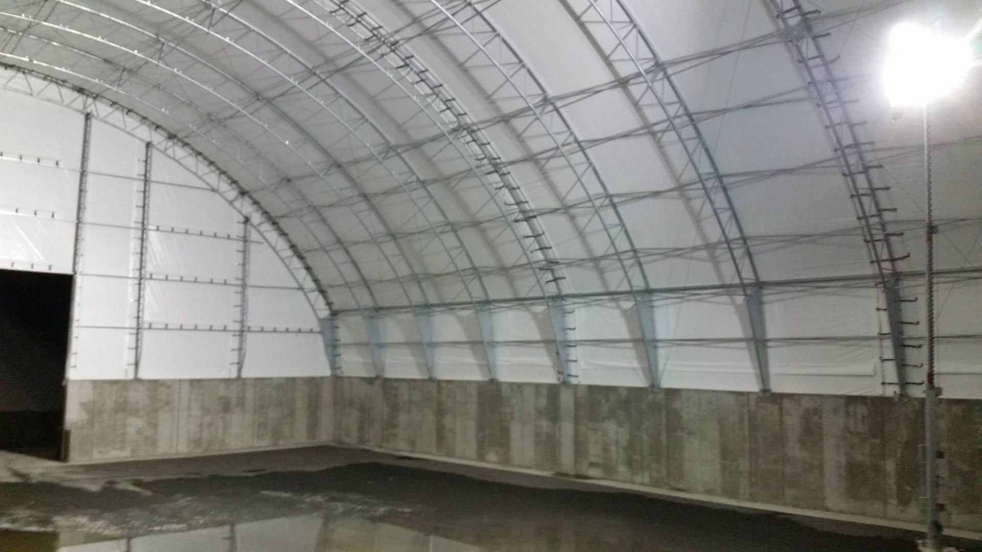 Interior of an arched industrial building with metal frame, white walls, and concrete base.