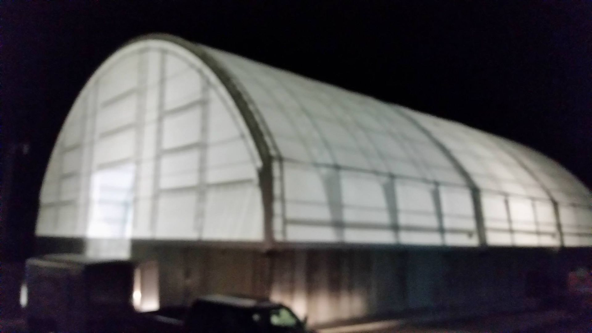 Arch-shaped, white-paneled building at night, with a dark base and a vehicle visible in the foreground.