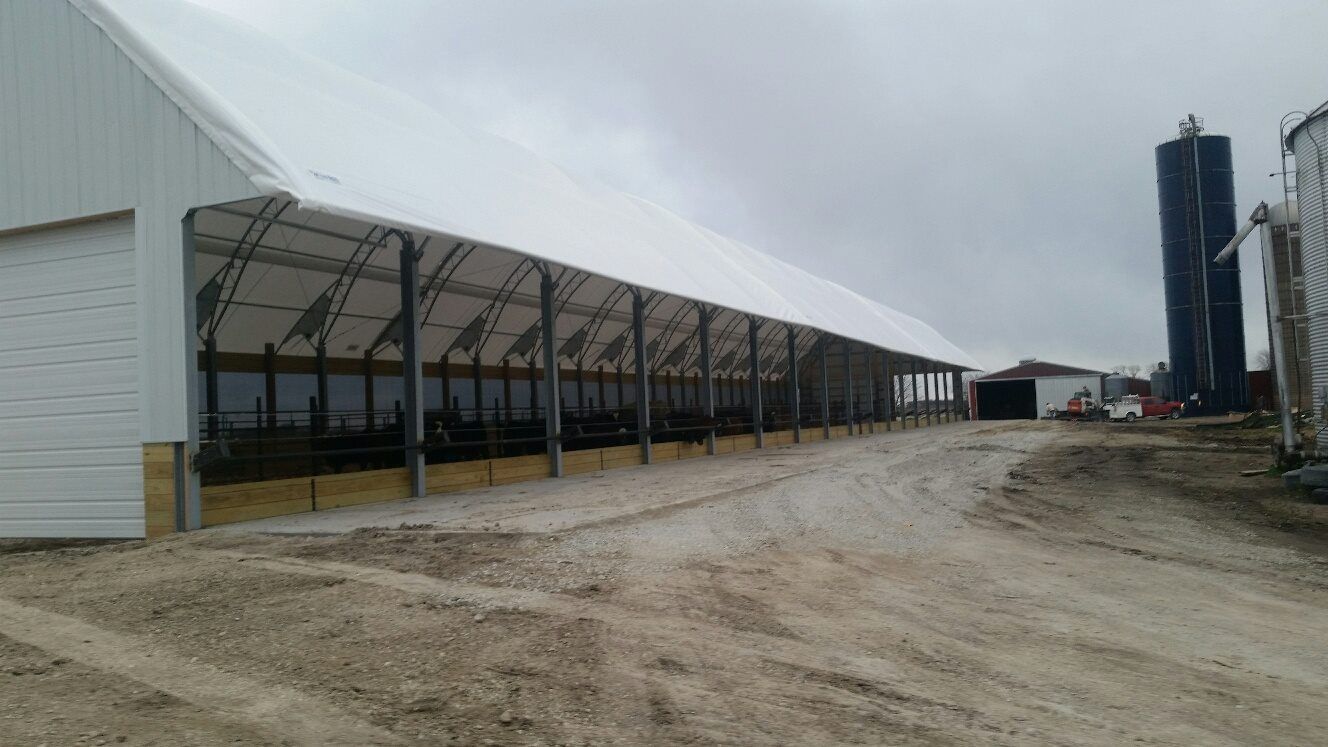 Long barn with white roof and open sides. Dirt road in front. Silo and other buildings visible.