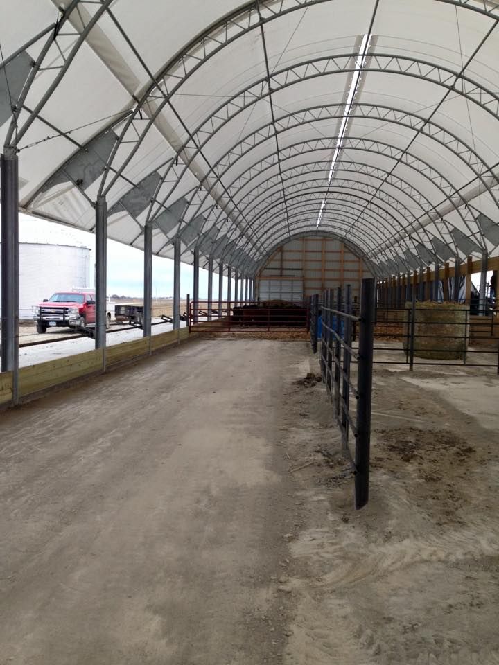 Inside of a long, white, arched-roof building with a dirt floor and metal fencing; a red pickup truck sits outside.