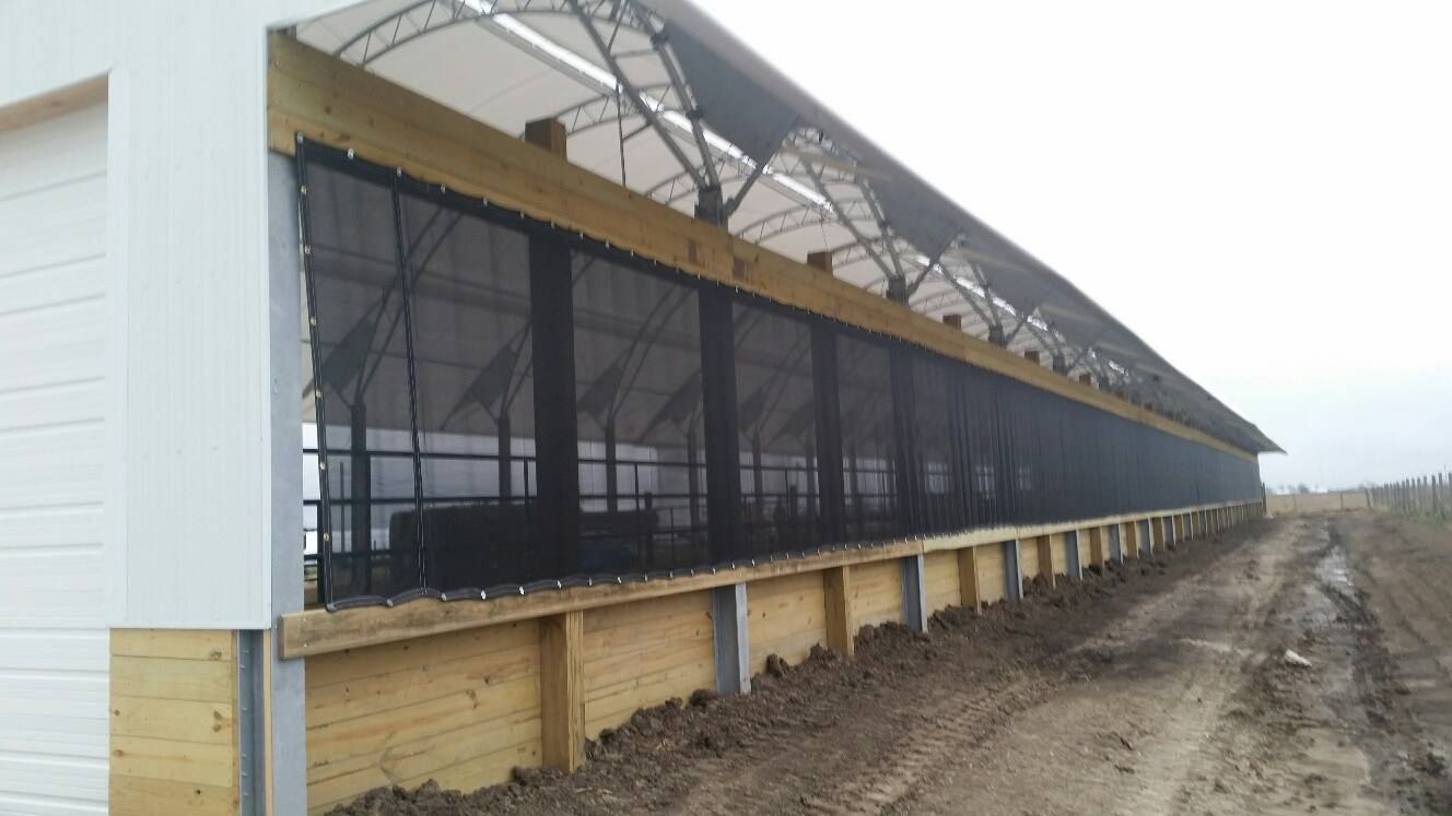 Long barn with black mesh sides, wooden frame, and clear roof panels. Dirt path in front.