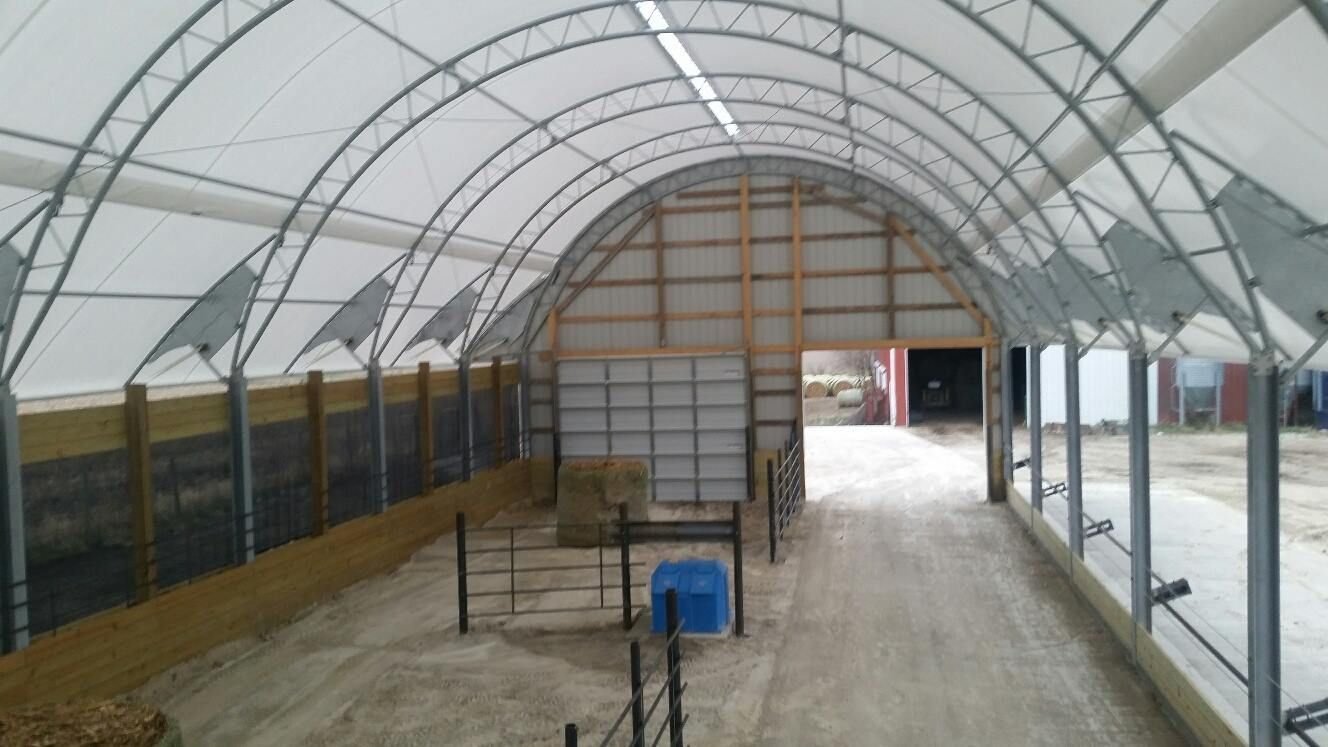 Interior of a barn with a curved metal frame roof, containing hay bales and fences.