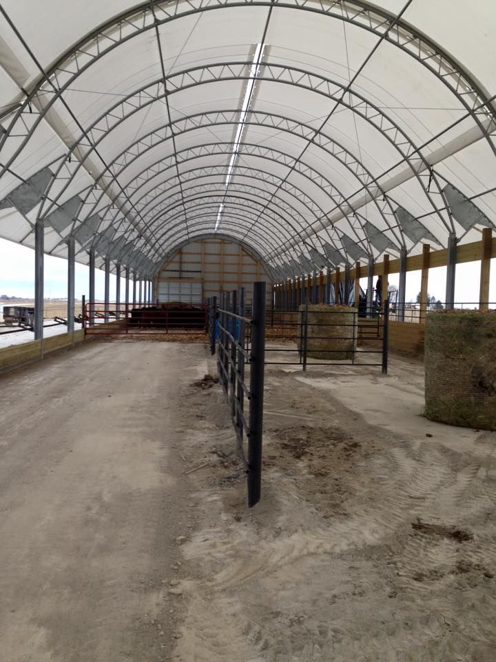 Open-air livestock shelter with arched roof and dirt floor; hay bales and fenced pens.