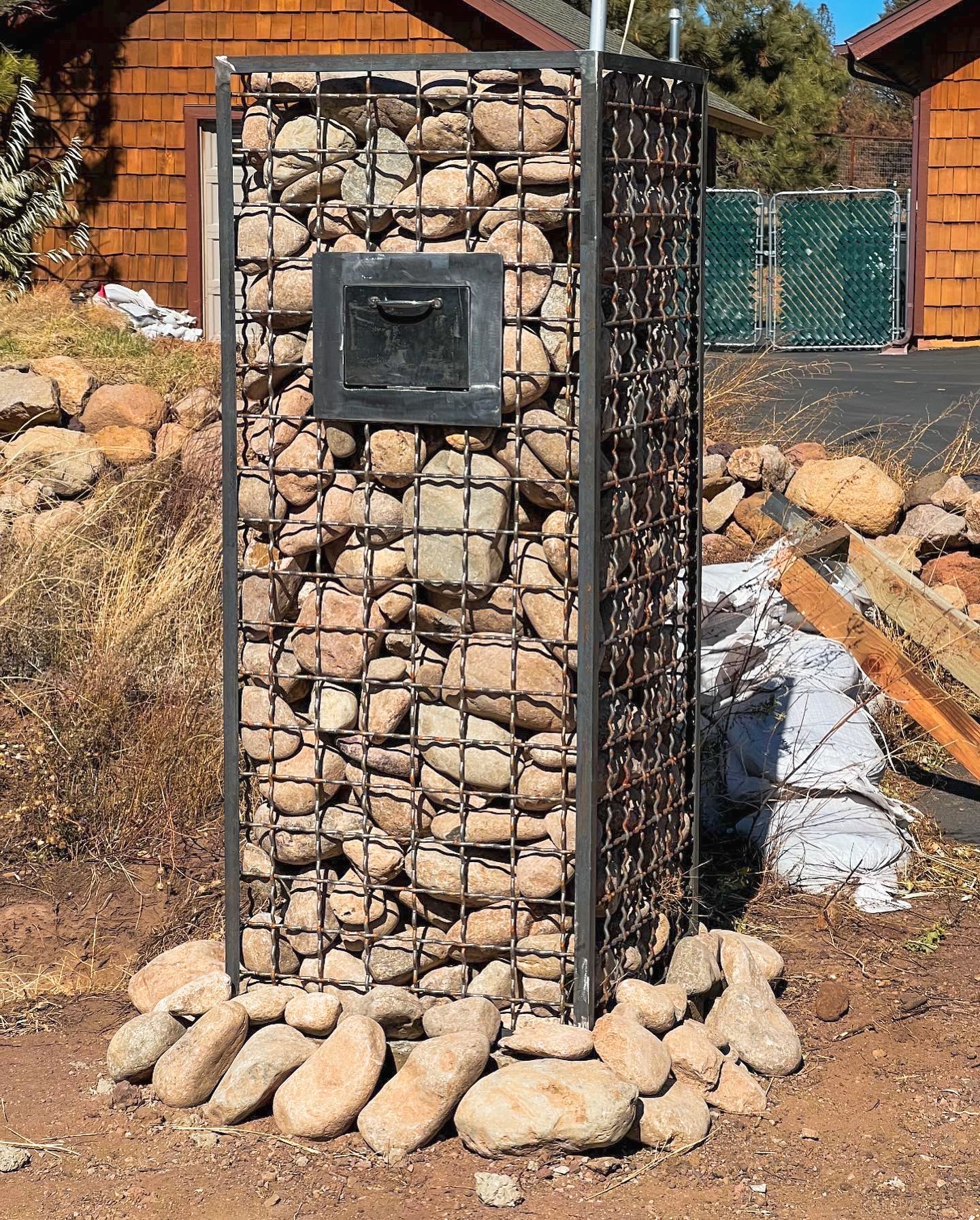 A cage filled with rocks is sitting in the dirt in front of a house.