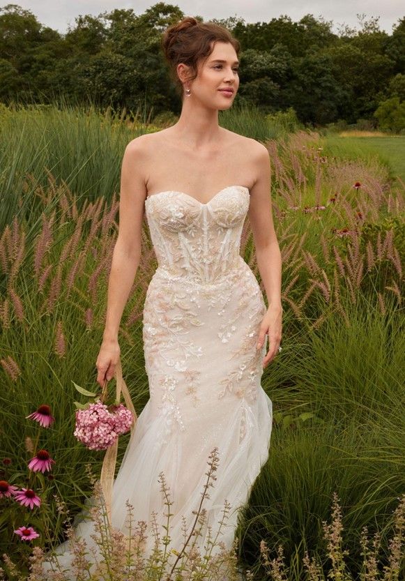 A woman in a wedding dress is standing in a field holding a basket of flowers - Salina, KS - Princess & Penguin Formalwear