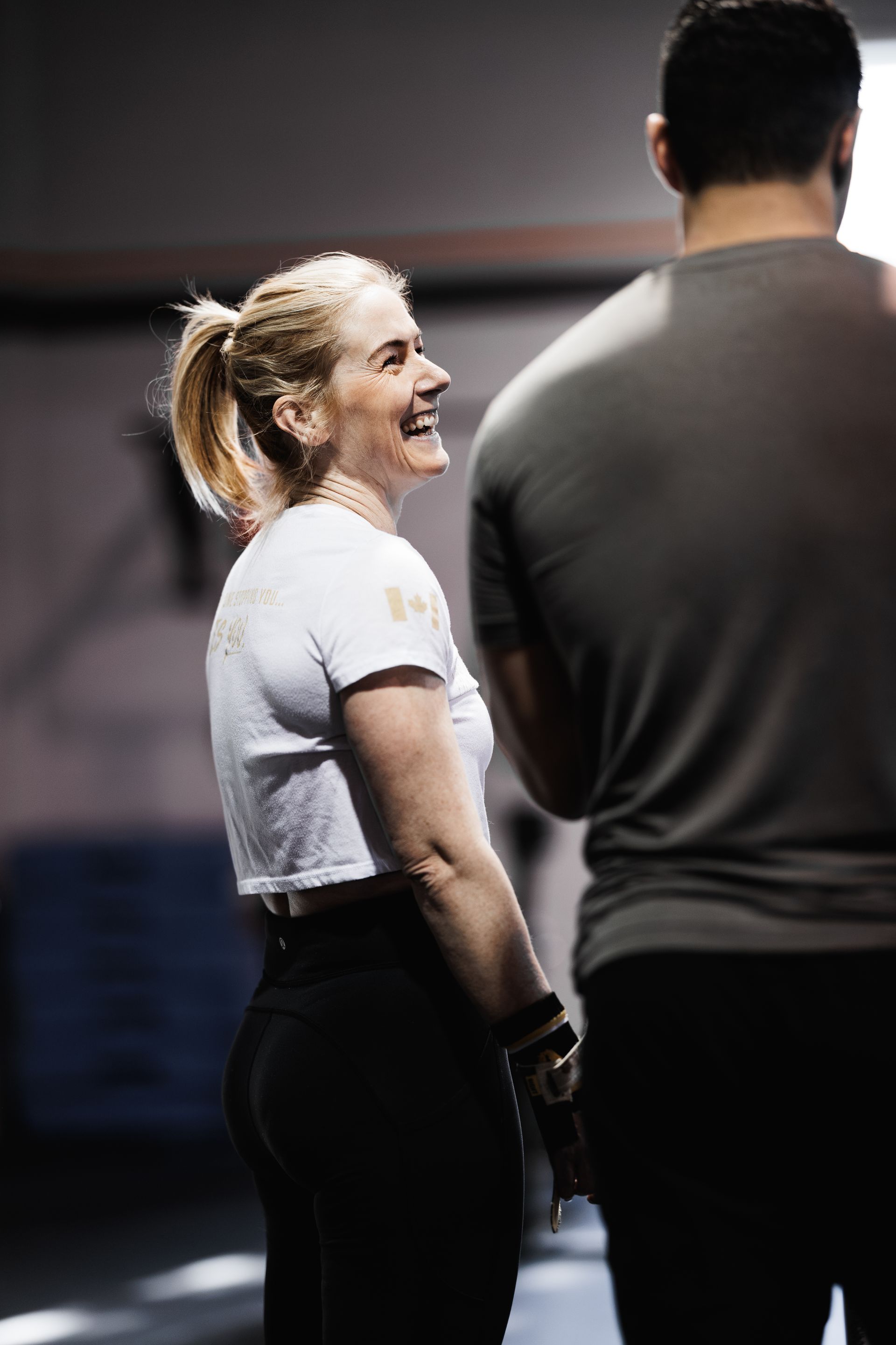 Woman smiles at person, wearing a cropped white top and black leggings, in a gym setting.