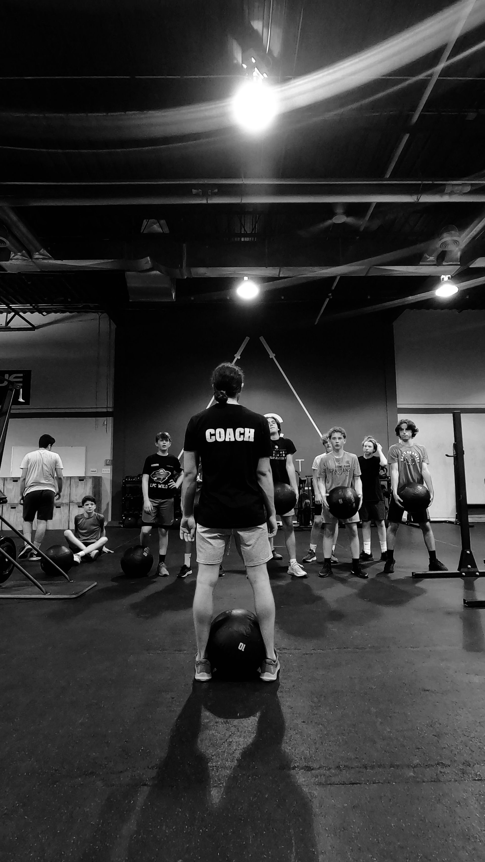 Coach facing group of young athletes in a gym; overhead light; black and white.