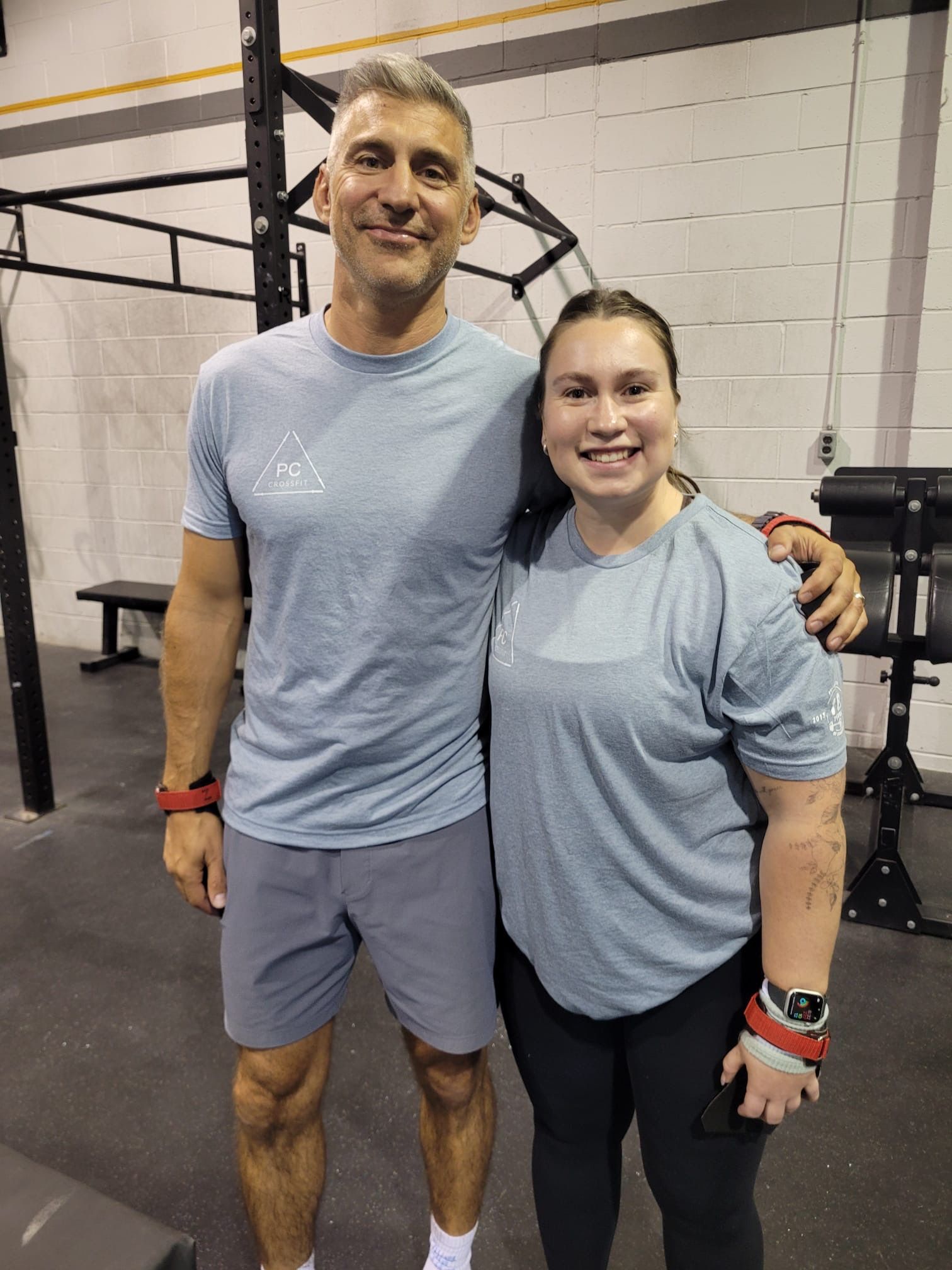 Two people pose for photo in a gym. Man has arm around woman. Both wear gray shirts and wrist wraps.