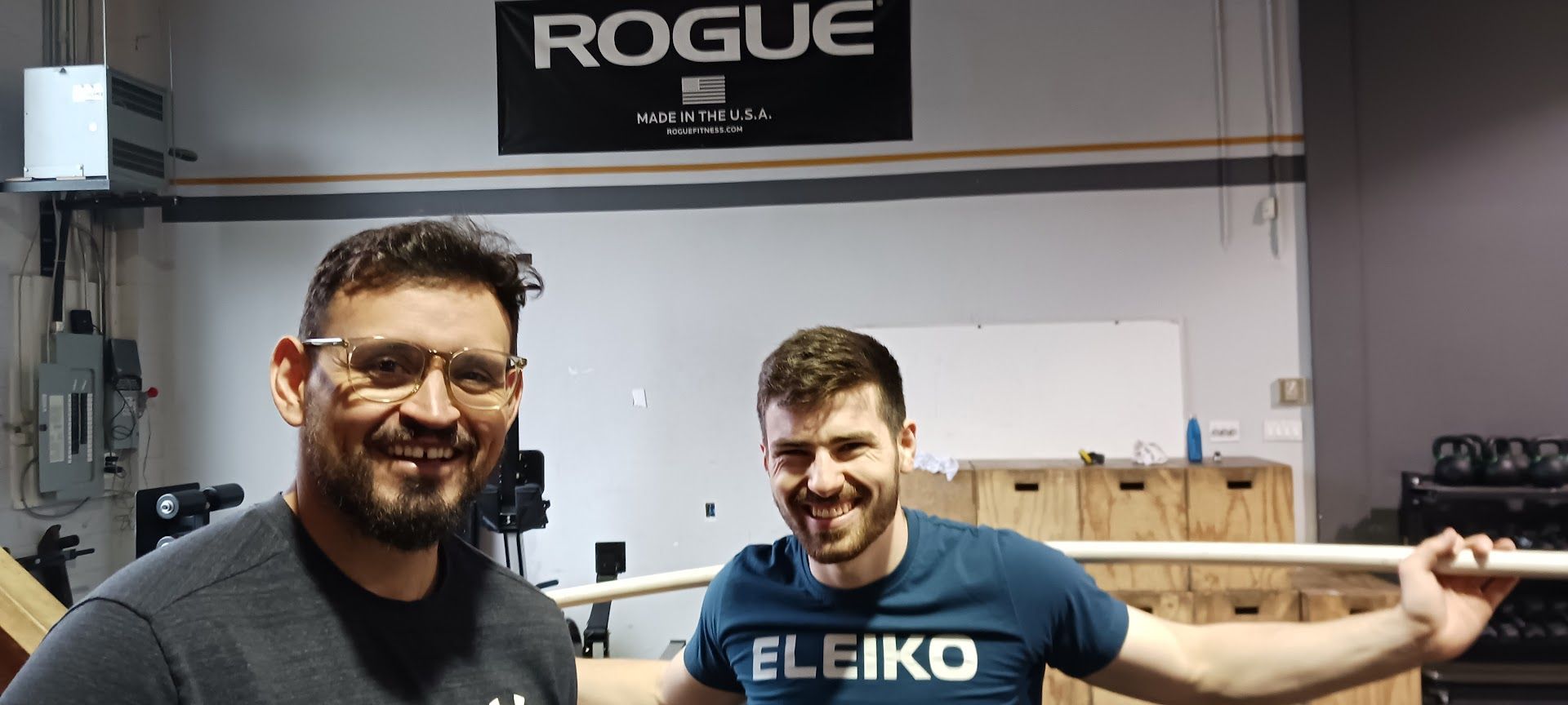 Two men smiling in a gym, one holding a barbell, 