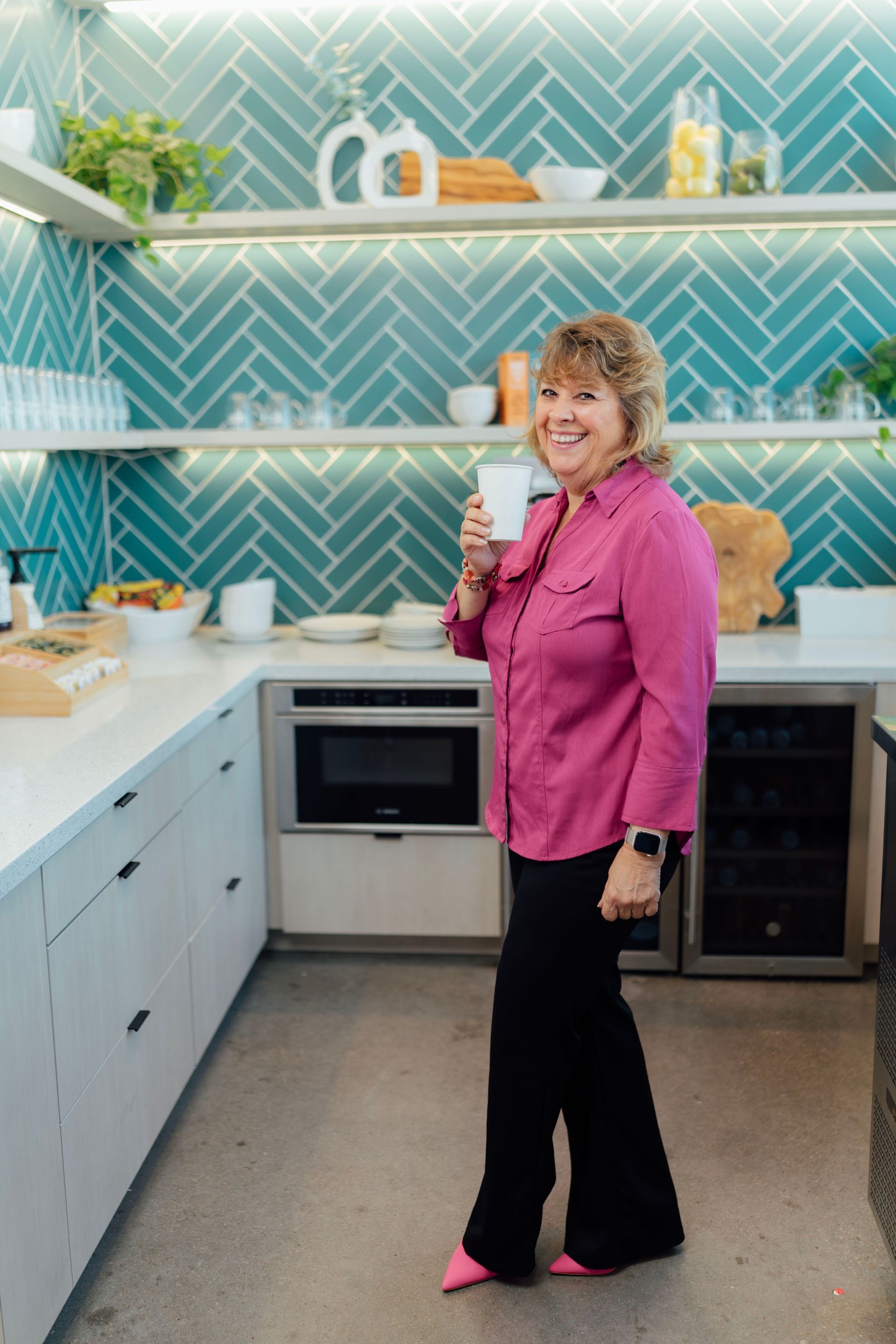 A woman in a pink shirt is standing in a kitchen holding a cup of coffee.