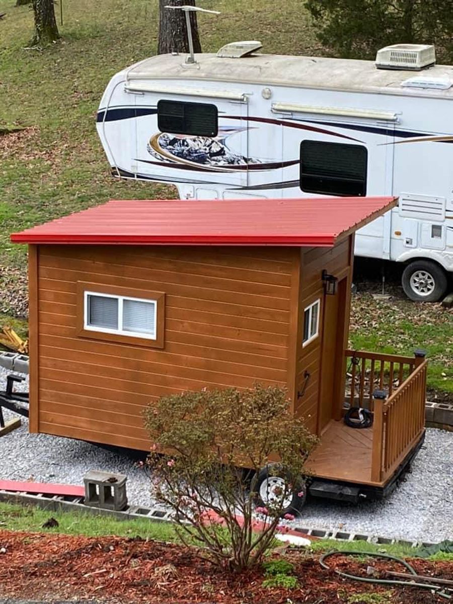Small wooden tiny house with red roof beside a white RV in a grassy campsite.