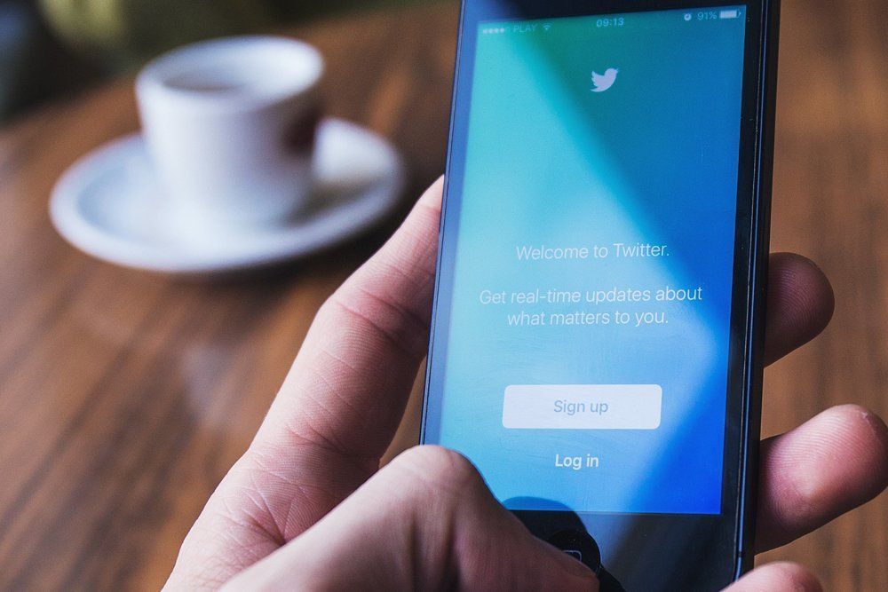 Person holding smartphone with Twitter login screen, blurred coffee cup in background.