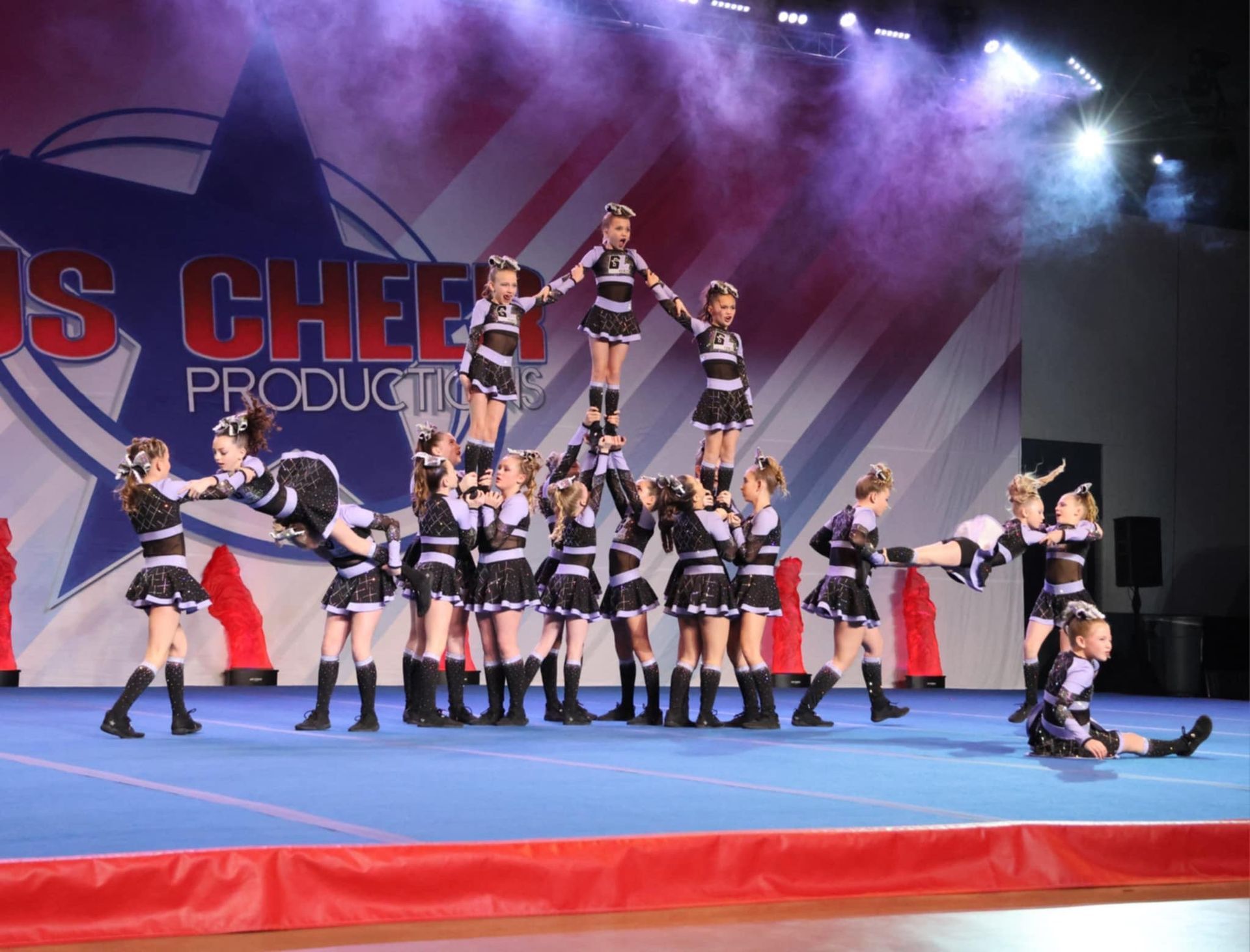 A group of cheerleaders are performing on a stage in front of a sign that says ' s cheer productions '