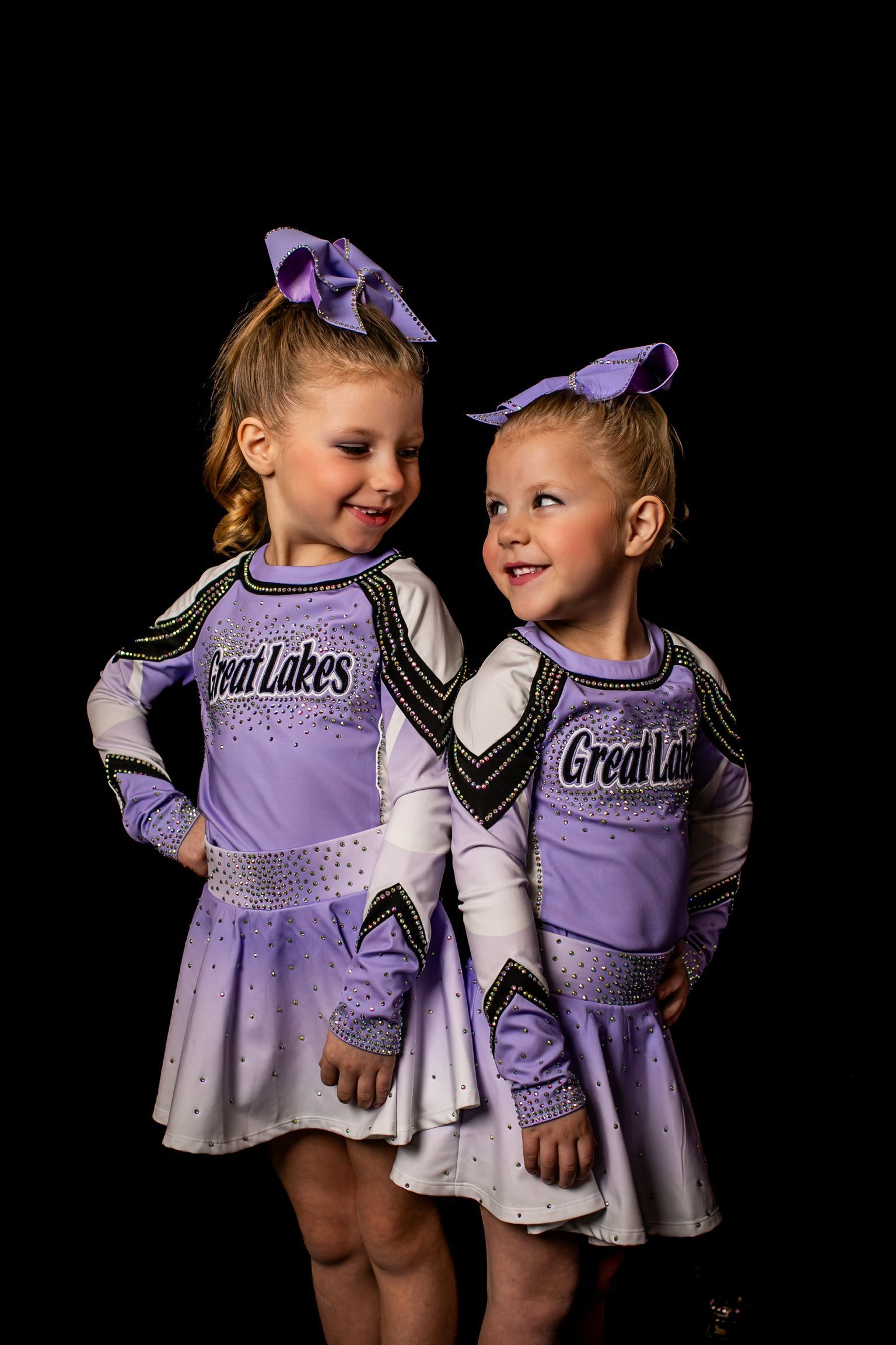 Two little girls in cheerleading uniforms are standing next to each other on a black background.