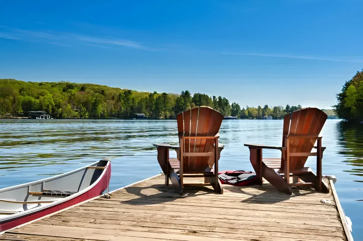Two wooden Adirondack chairs sit on a dock beside a canoe, overlooking a lake with trees. — AT My Place Counselling and Psychotherapy in Neutral Bay, NSW