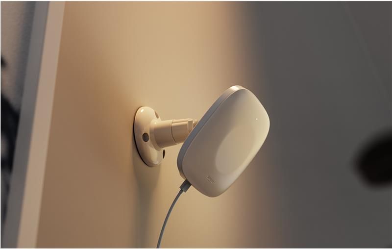 A wall-mounted, white contactless monitoring device designed to collect patient vital information. It is attached to a beige wall using an adjustable bracket and connected via a power cable. The sleek, rounded design allows for unobtrusive, passive monitoring in a clinical or care facility setting.
