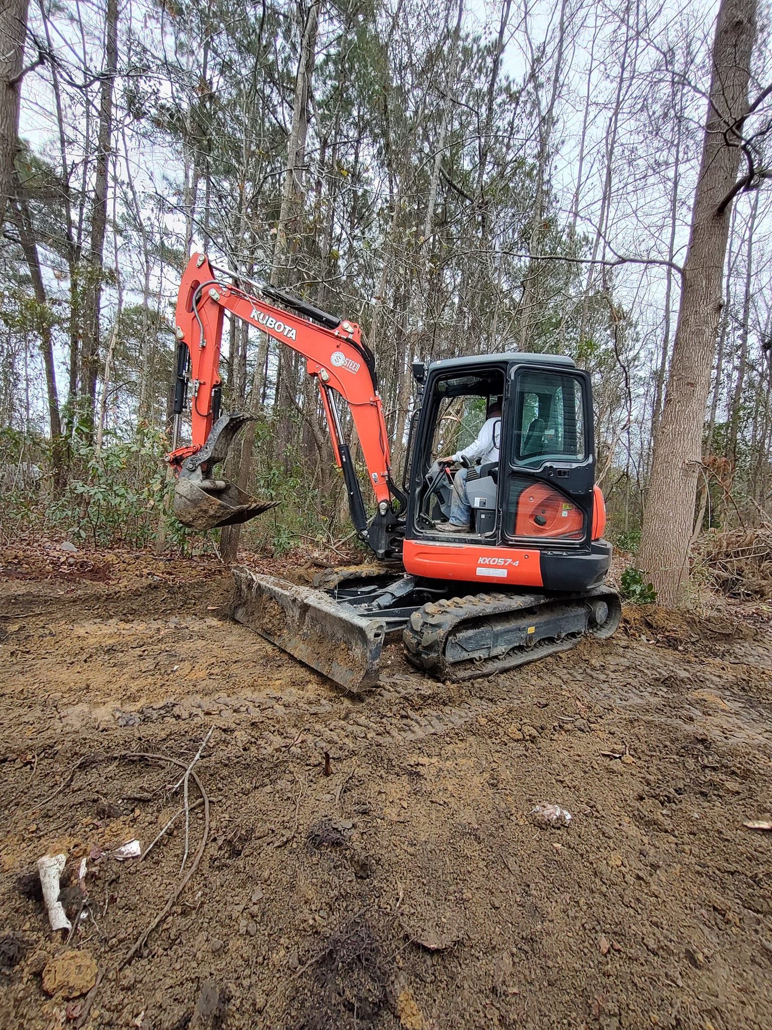 A red and black excavator is digging in the dirt in the woods.