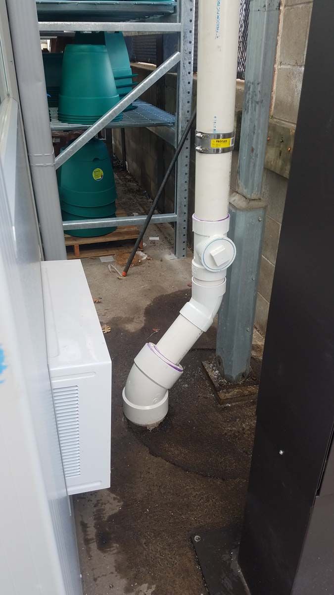 White PVC pipe bends down from a vertical drain near a building, next to a white appliance.