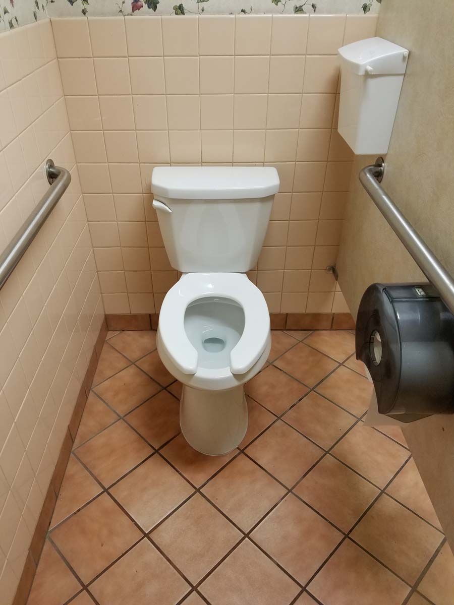 A public restroom with a white toilet, tan walls, and brown tile floor. Handrails are present.