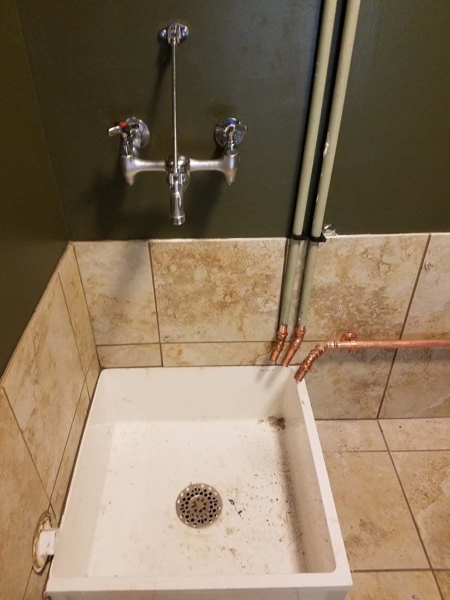 A wall-mounted faucet over a white sink in a tiled corner, with exposed copper pipes.