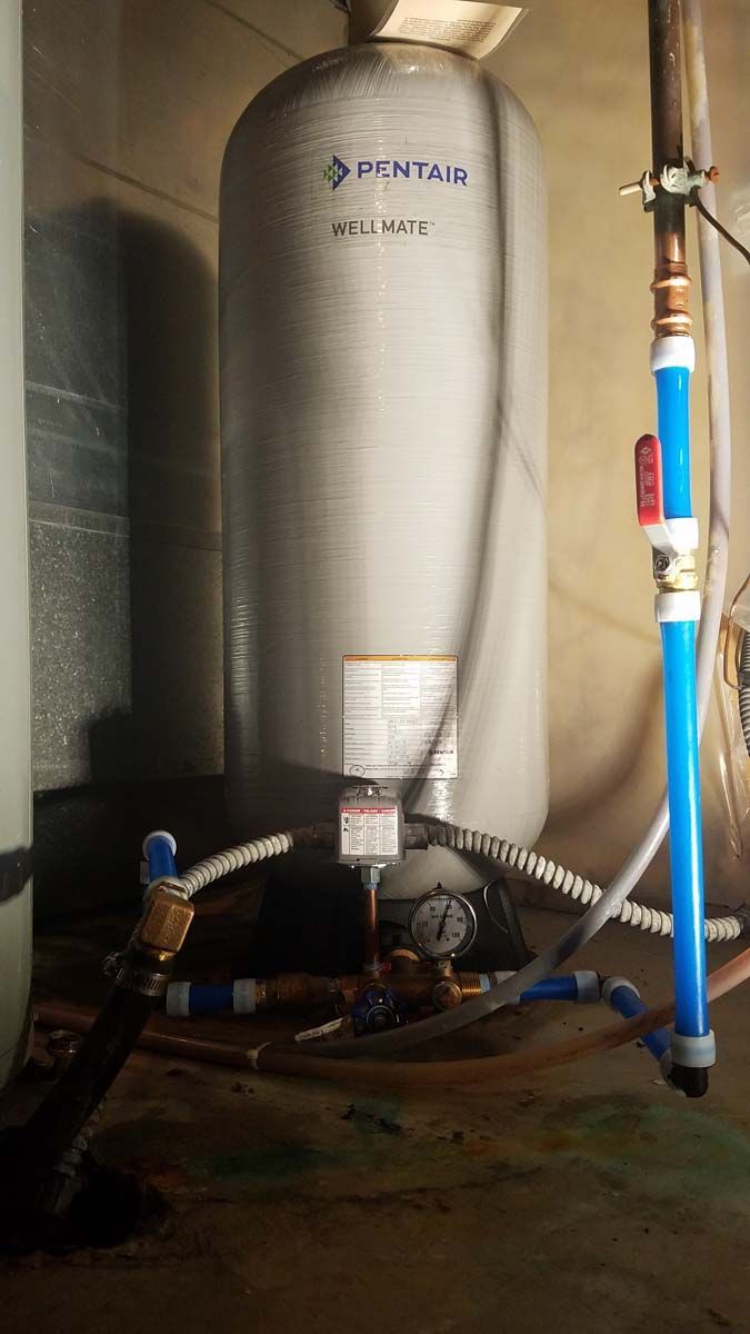 Water pressure tank in a dimly lit basement. Blue and copper pipes connected to the tank and surrounding equipment.