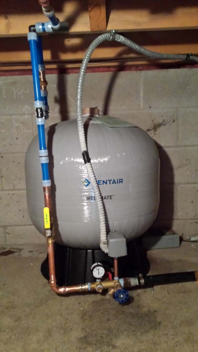 A water pressure tank with copper and blue pipes in a basement against a cinder block wall.