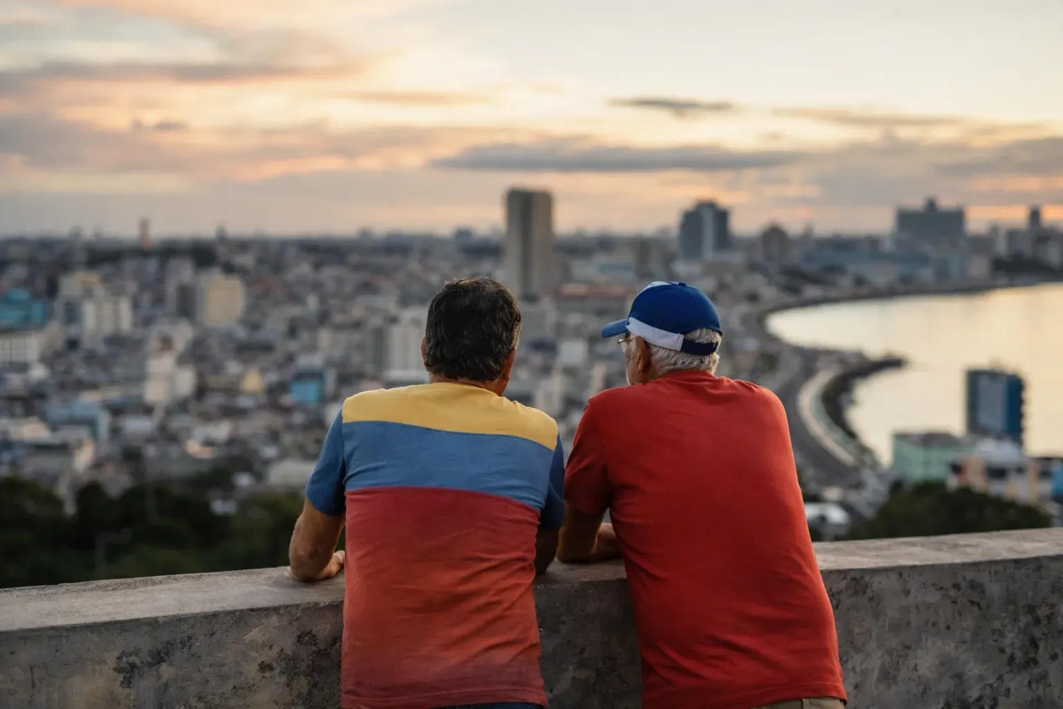 Two people stand at a stone ledge overlooking a coastal city during sunset, with a curved shoreline visible.