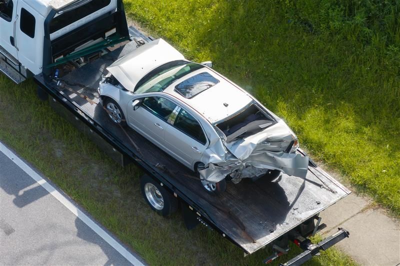 A severely damaged silver car on a flatbed tow truck, roadside.
