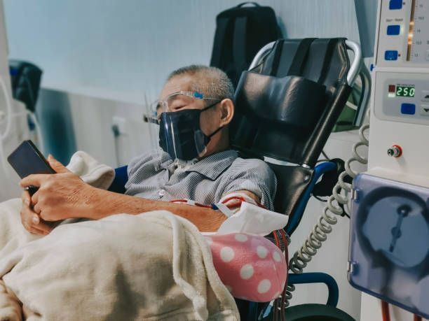 An elderly man wearing a mask sits in a dialysis chair, looking at his phone in a hospital setting.