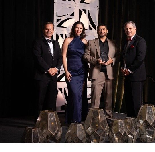 Four people on stage, holding an award, dressed in formal attire, at an event with decorative background.