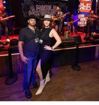 A couple poses at a live music venue, stage in background. Man wears hat, woman in dress, both smiling.