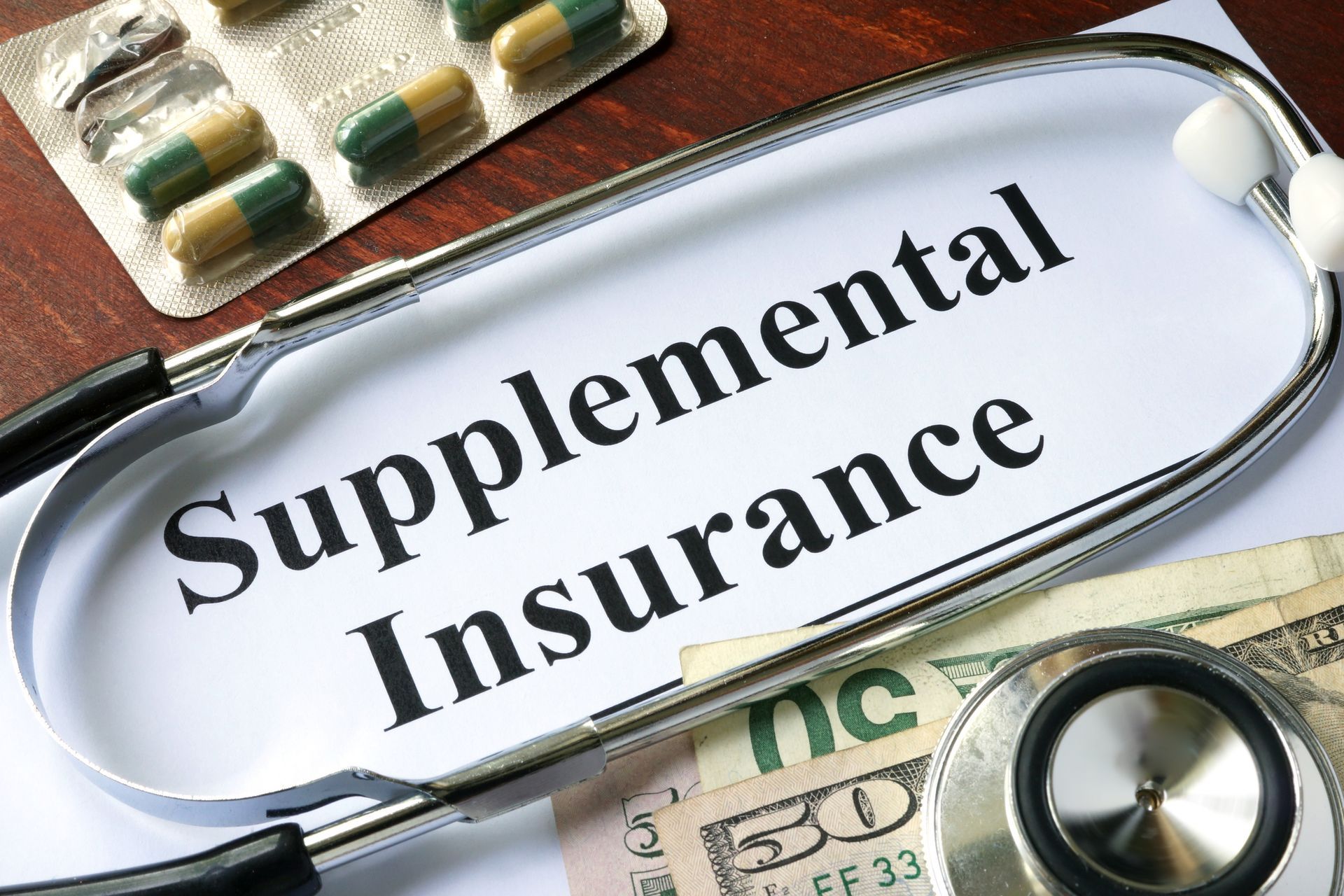 Supplemental insurance written on a paper with a stethoscope and dollar bills on top and pills next to it.