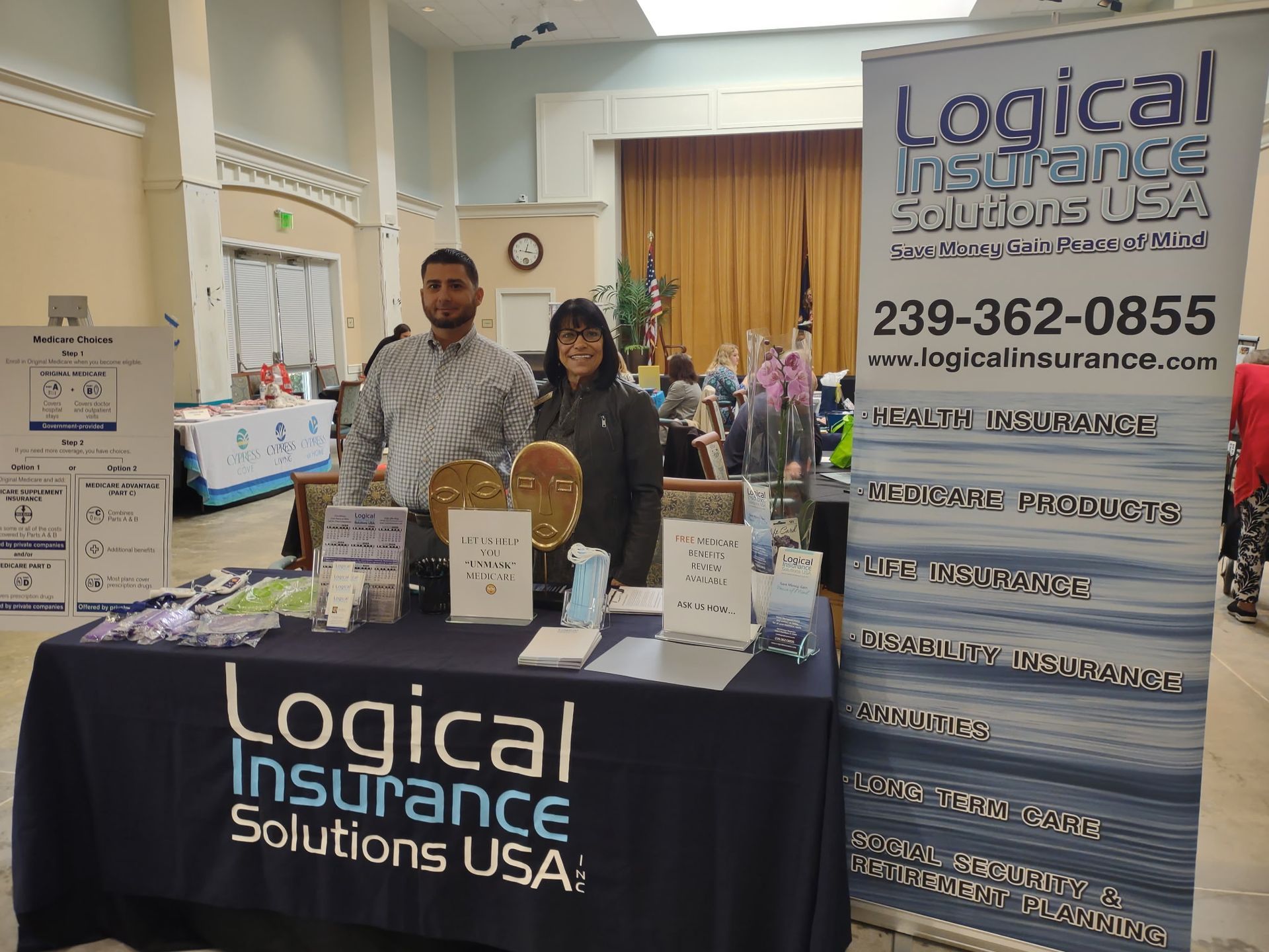 Two people at a Logical Insurance Solutions USA booth. Navy table, sign, banner; Florida phone, web address.