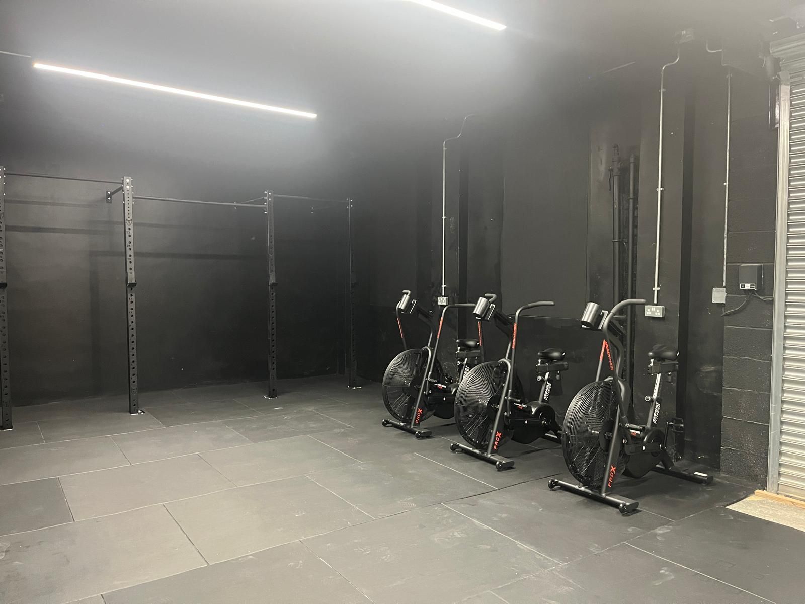 A minimalist gym with black walls and floors, featuring a pull-up rig on the left and three stationary air bikes in a row.