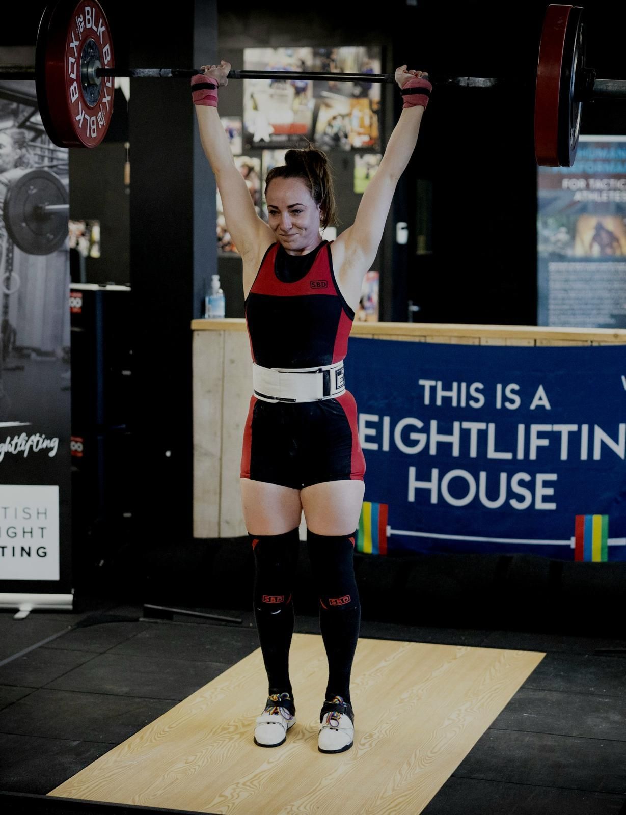 A weightlifter in a red and black singlet successfully completes a barbell overhead lift in a gym.