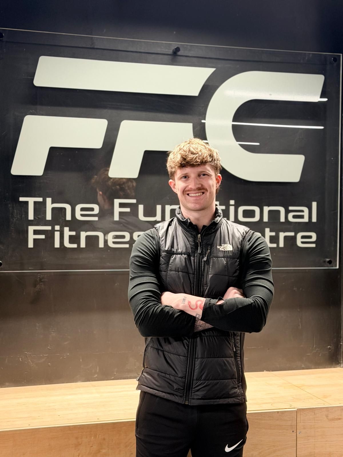 A person stands with arms crossed in front of a sign that reads 