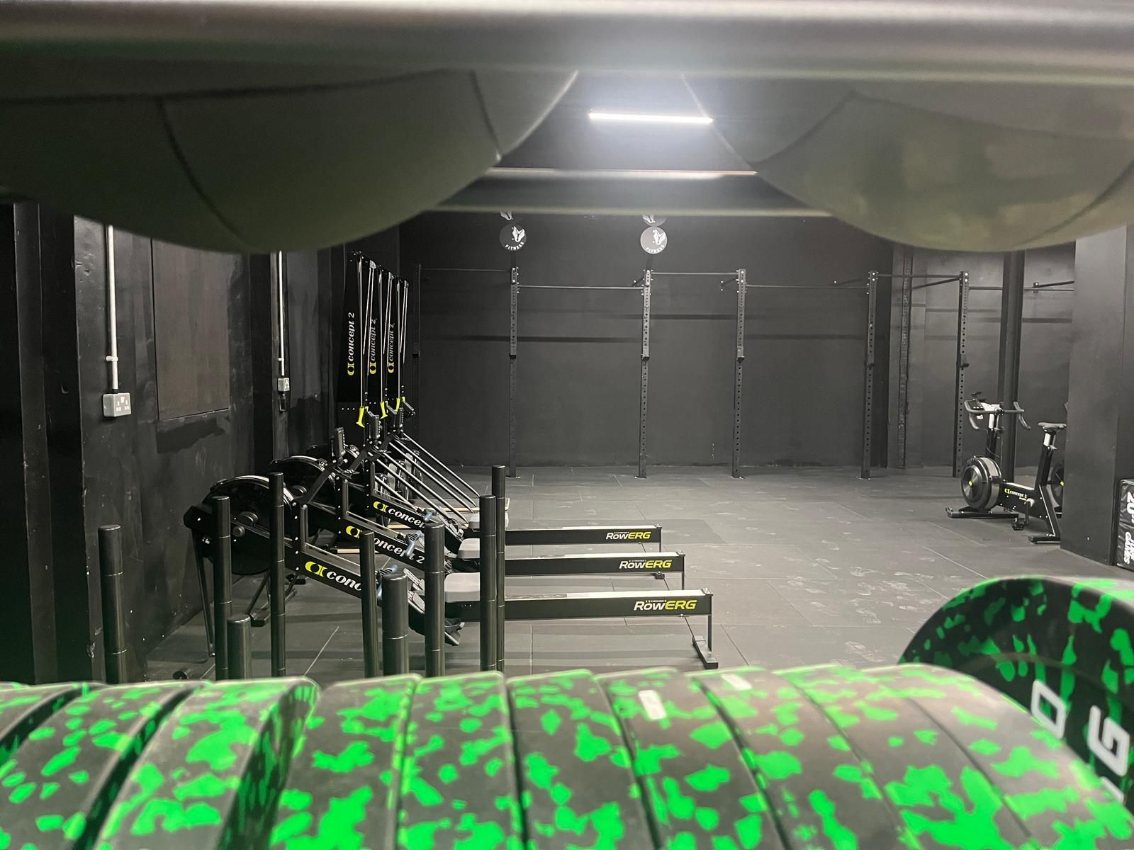 A dimly lit gym interior featuring rows of rowing machines, black walls, and green-patterned weight plates in the foreground.