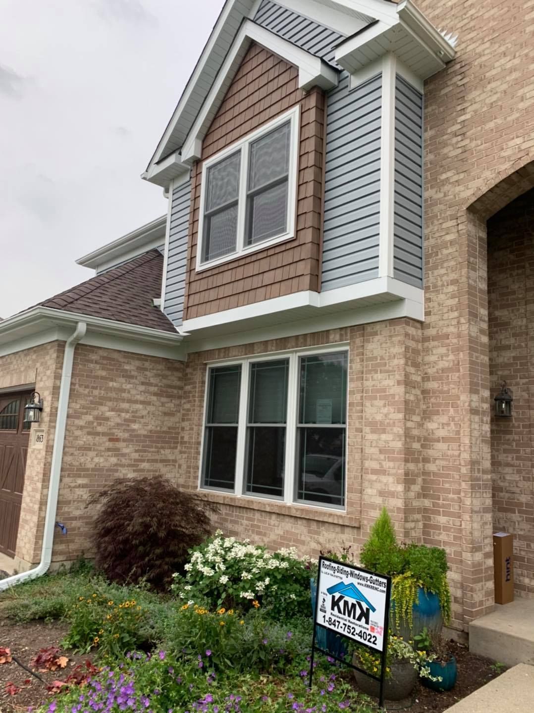 A beige brick home featuring a two-story bay window with brown siding, gray accents, and a business sign in the front yard.
