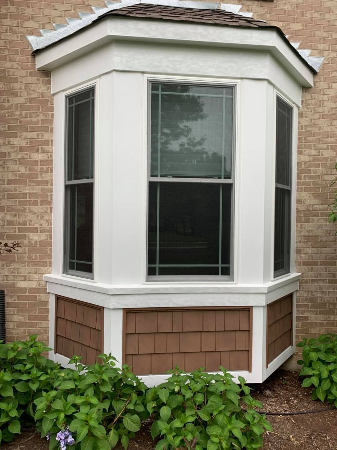 A white, three-sided bay window set into a brick exterior wall, featuring brown cedar shake siding beneath the glass.