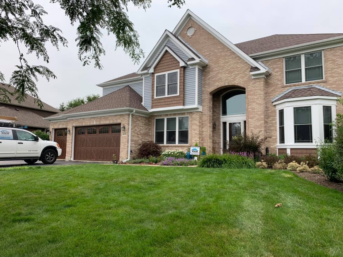 A large, two-story tan brick suburban house with a brown roof, attached garage, and a green lawn under a cloudy sky.