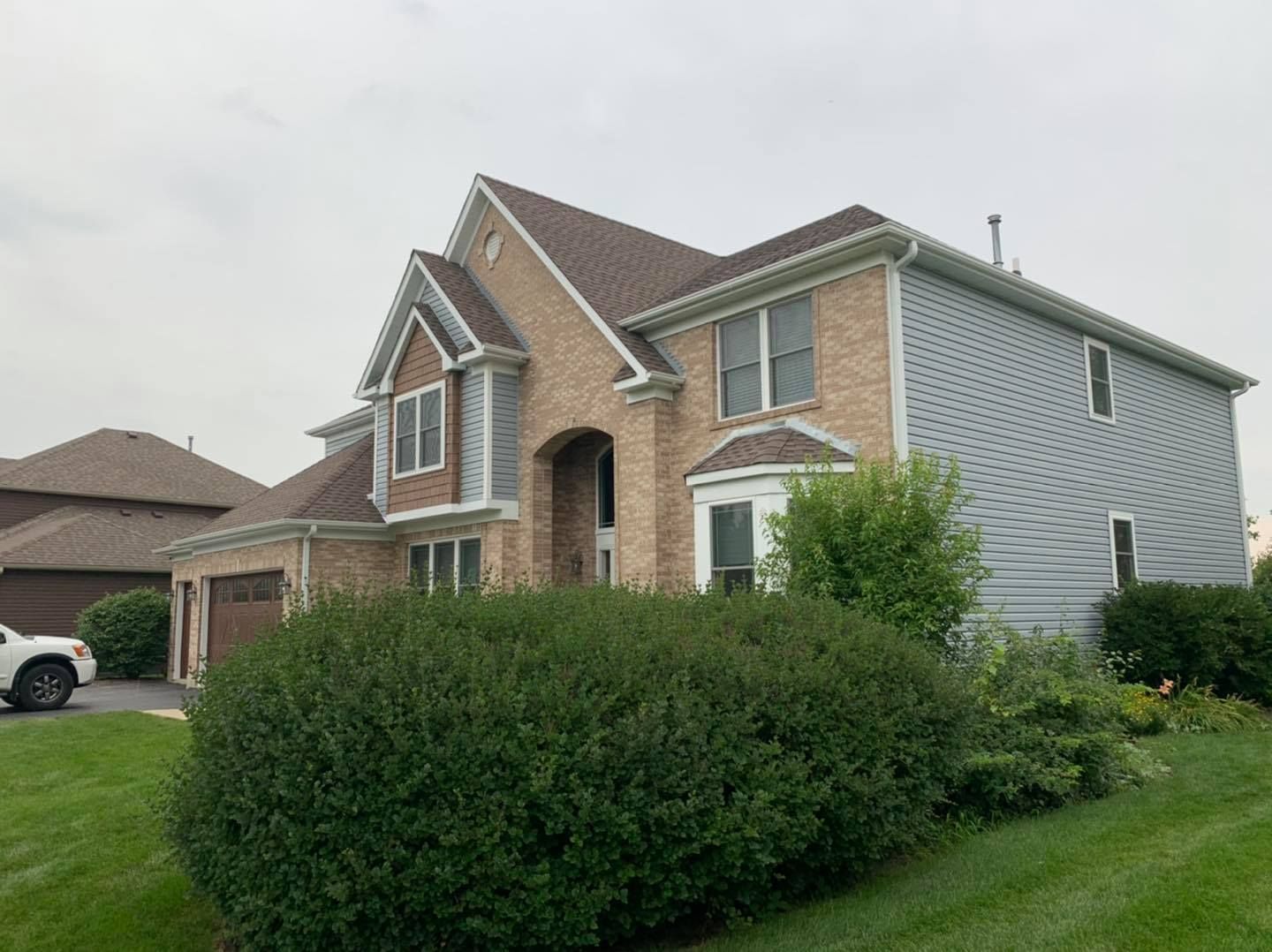 A two-story house with beige brick, gray siding, a brown roof, and a large rounded hedge in the front yard.