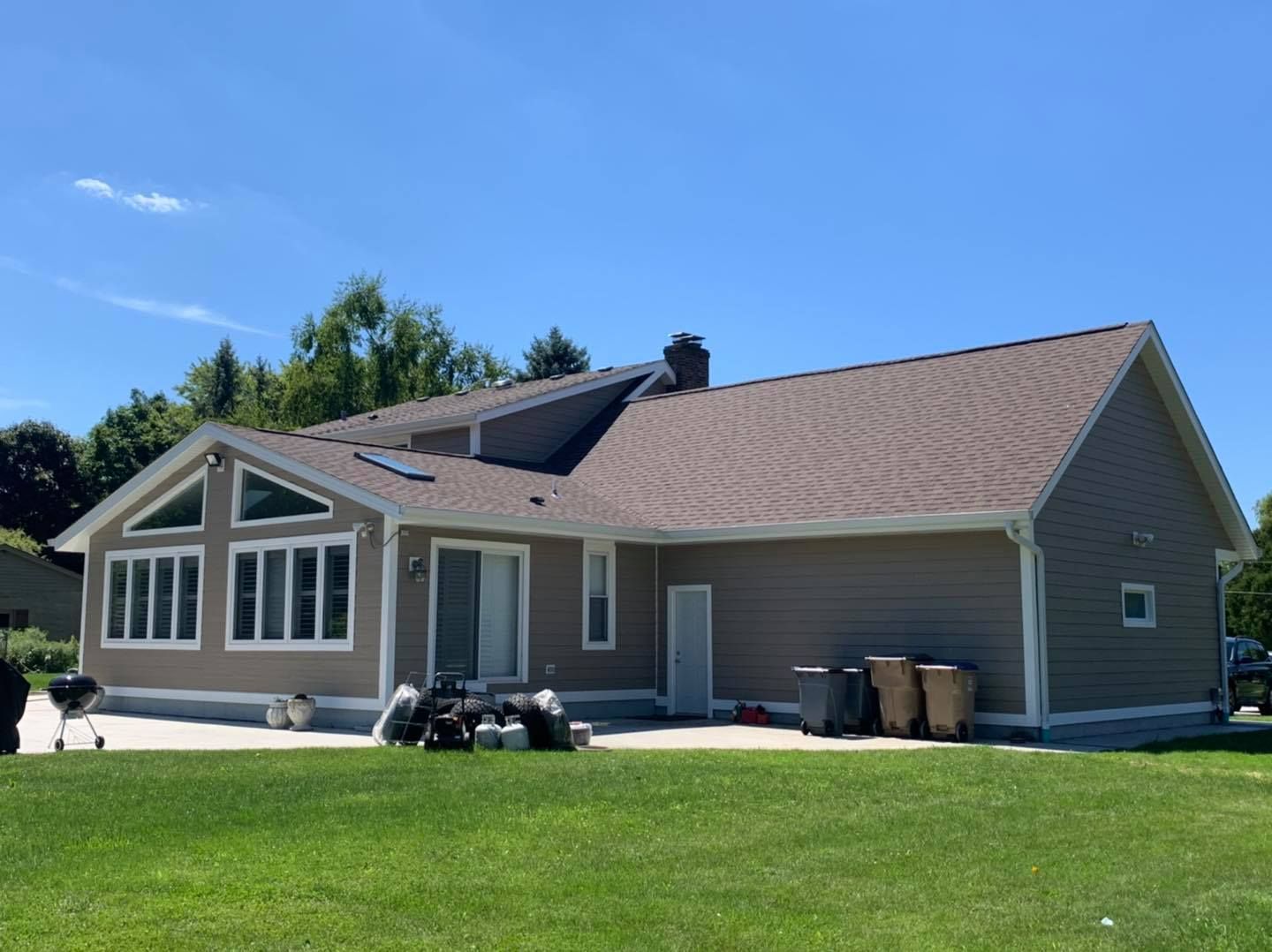 A tan house with a sunroom, gray shingles, and white trim sits on a green lawn under a clear blue sky.