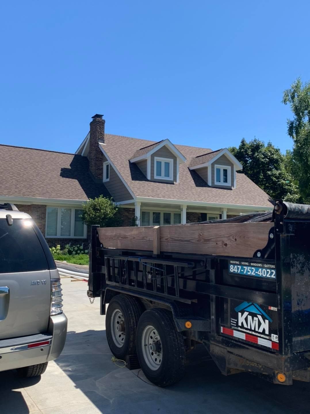 A black dump trailer for KMX Roofing parked in a residential driveway in front of a two-story home with a new roof.