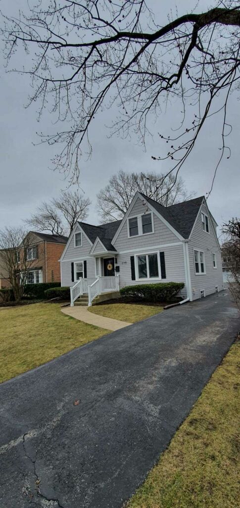 A single-story gray house with a dark roof and white trim, featuring a paved driveway and lawn under a cloudy sky.