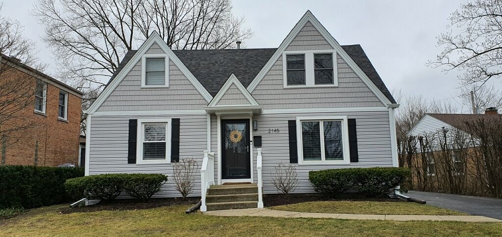 A single-story, light-gray house with two front gables, a dark front door, and black shutters, set behind a small lawn.