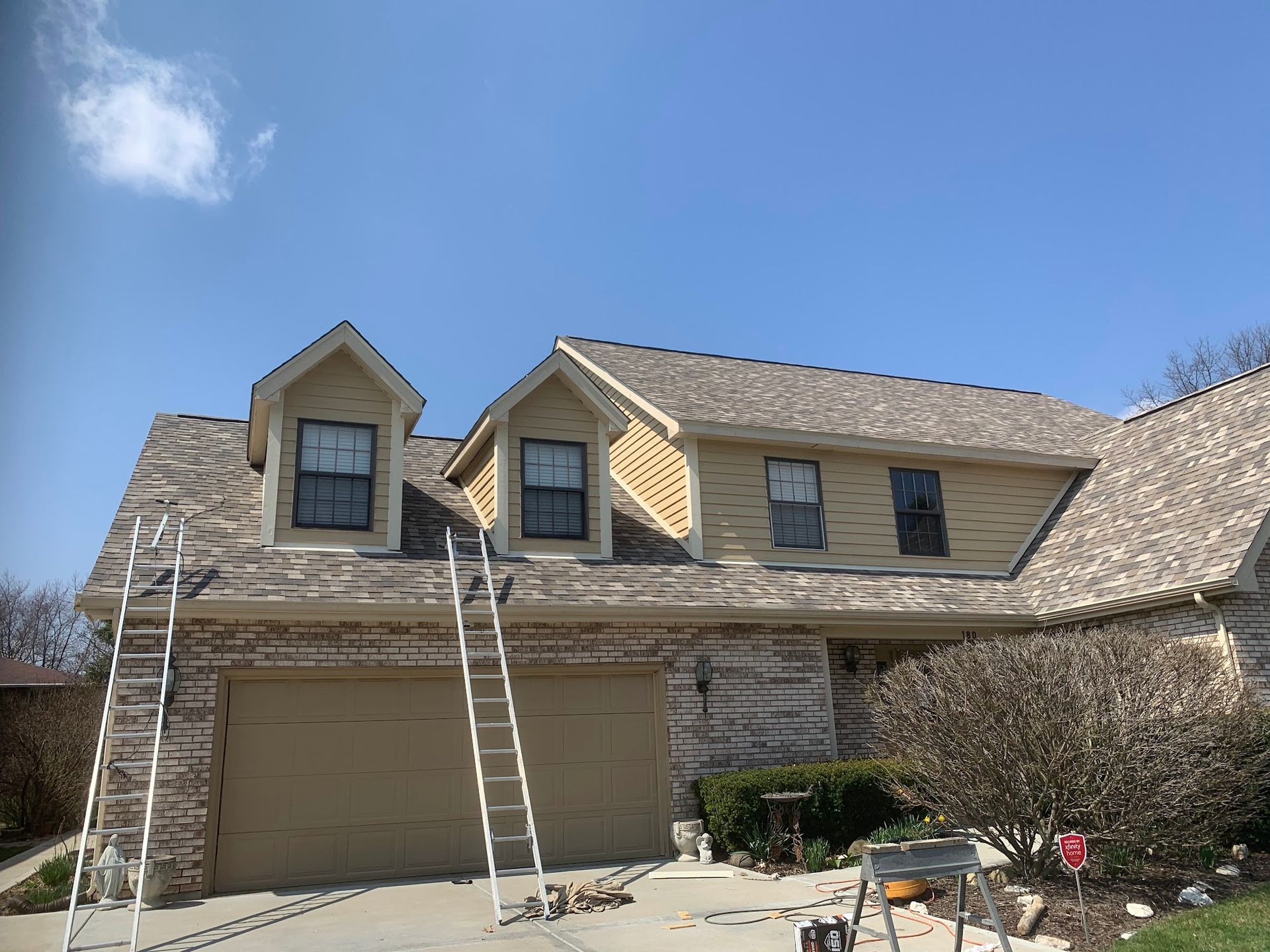 A two-story house with a brick and tan siding exterior, featuring two dormer windows and two ladders leaning on the roof.