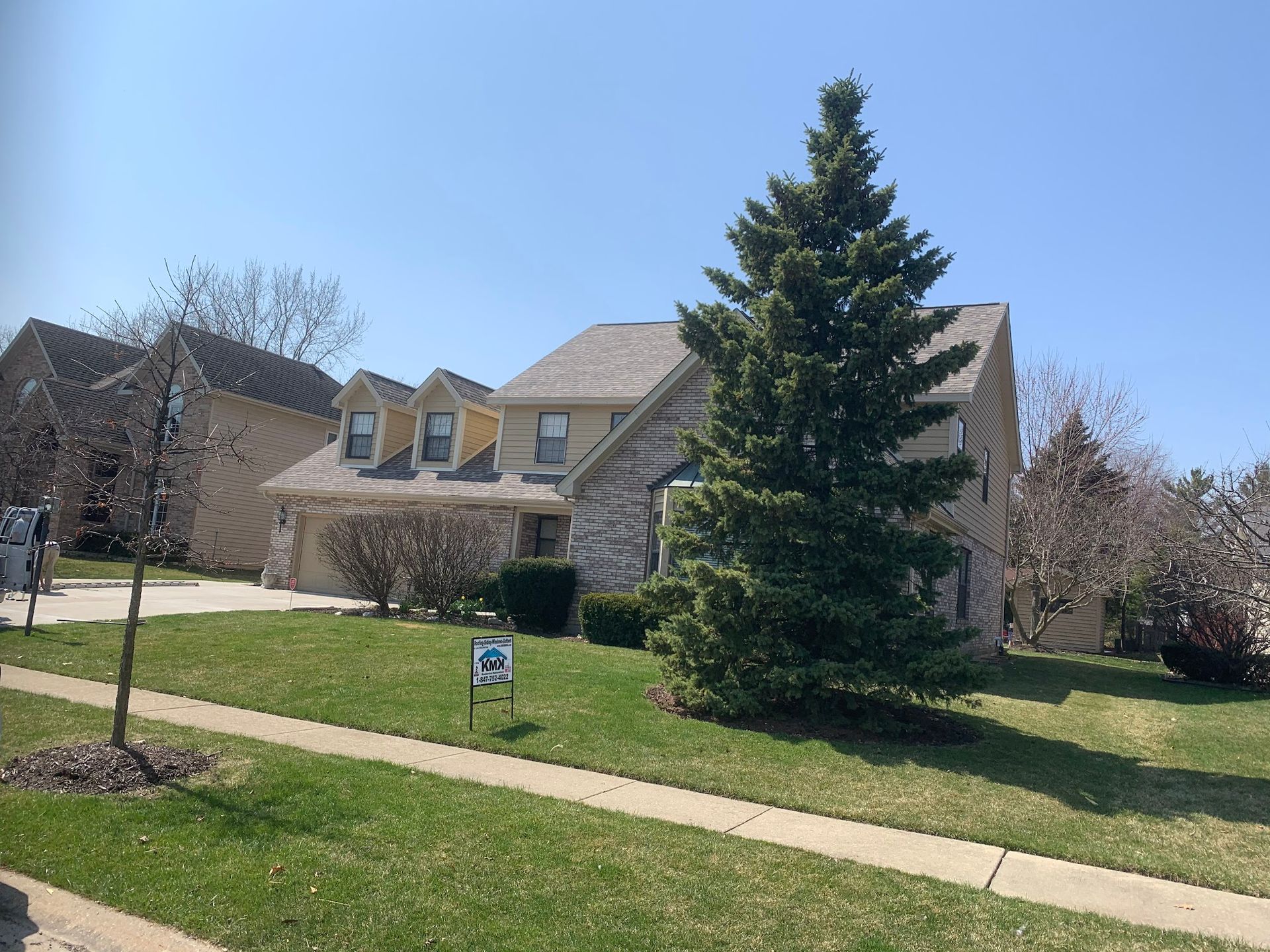 A two-story tan brick house with dormer windows and a tall evergreen tree in the front yard under a clear blue sky.