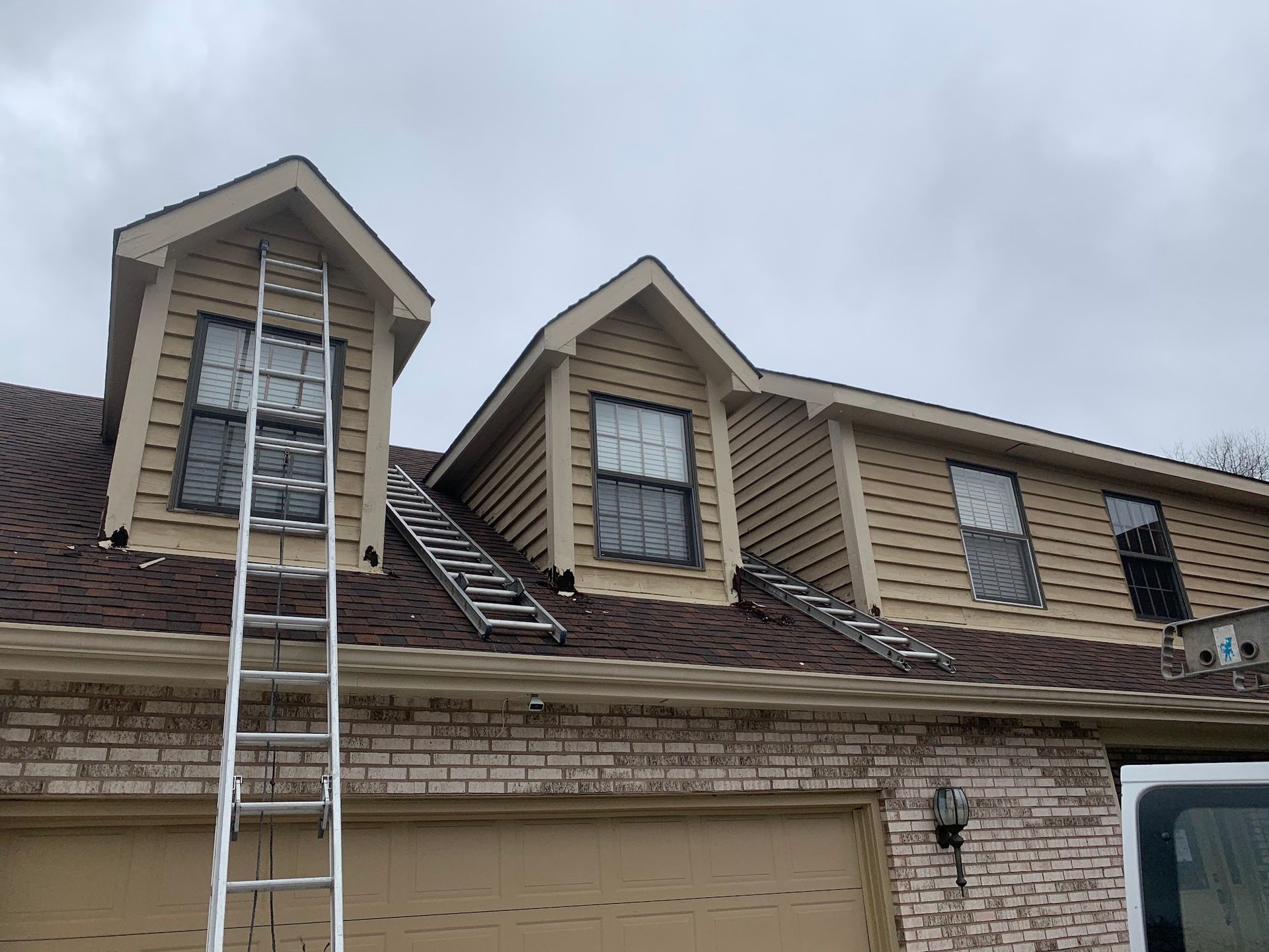 Ladders lean against the tan siding and brown shingled roof of a house with multiple dormer windows under a cloudy sky.